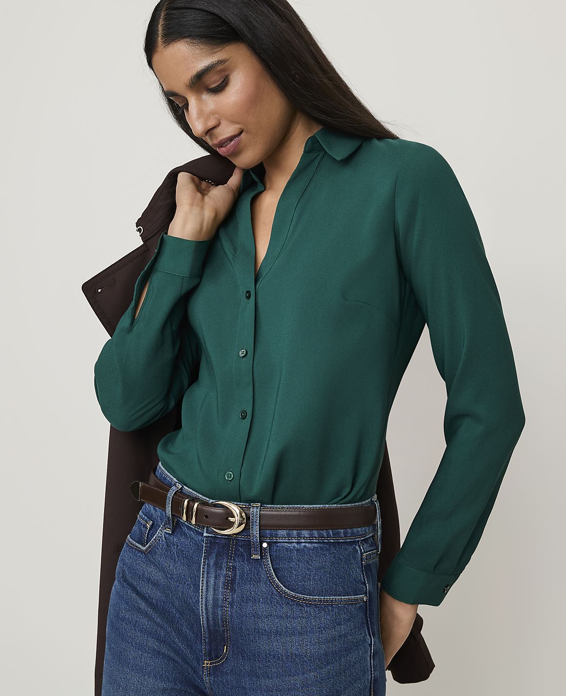 A woman in a green blouse and jeans stands against a plain background, with a brown jacket draped over her shoulder.