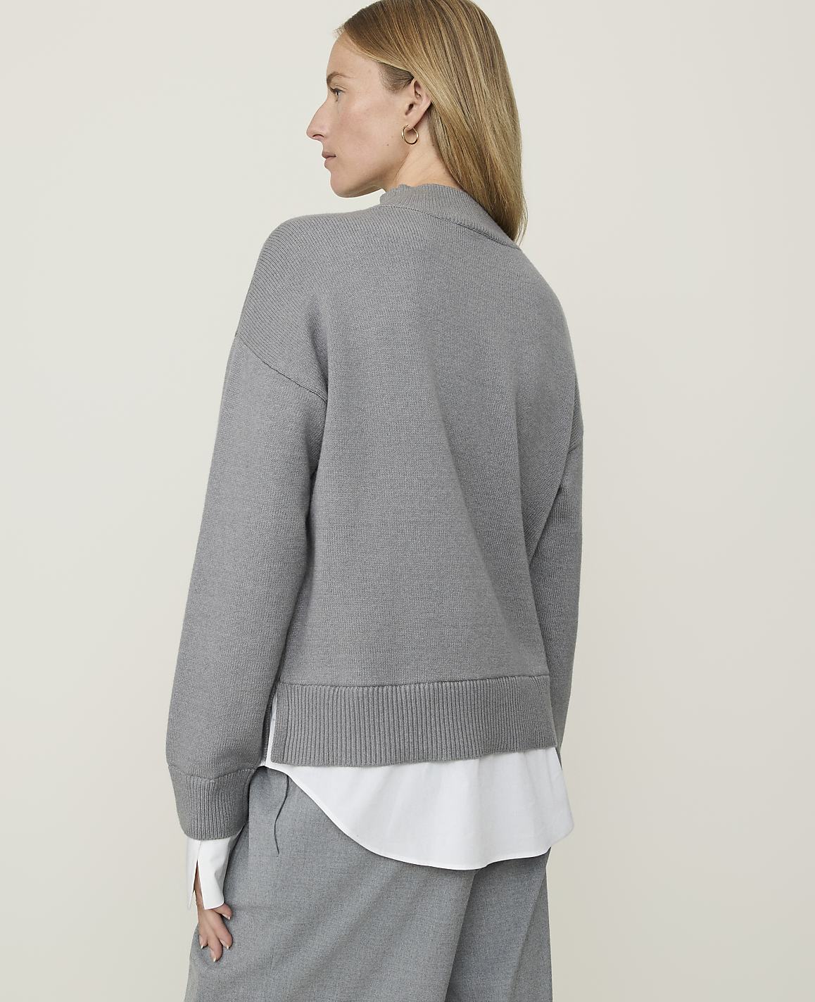 A woman in a grey sweater and white shirt stands against a plain, light grey background.