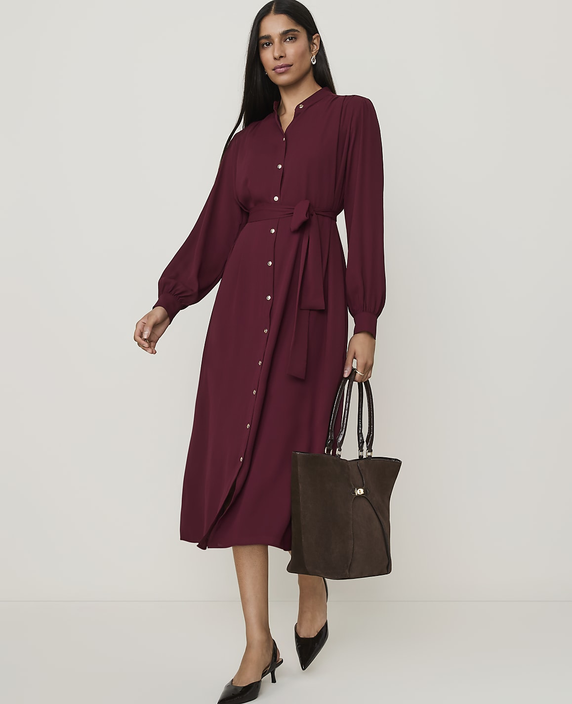 A woman in a burgundy dress and black heels stands with a brown tote bag against a plain background.