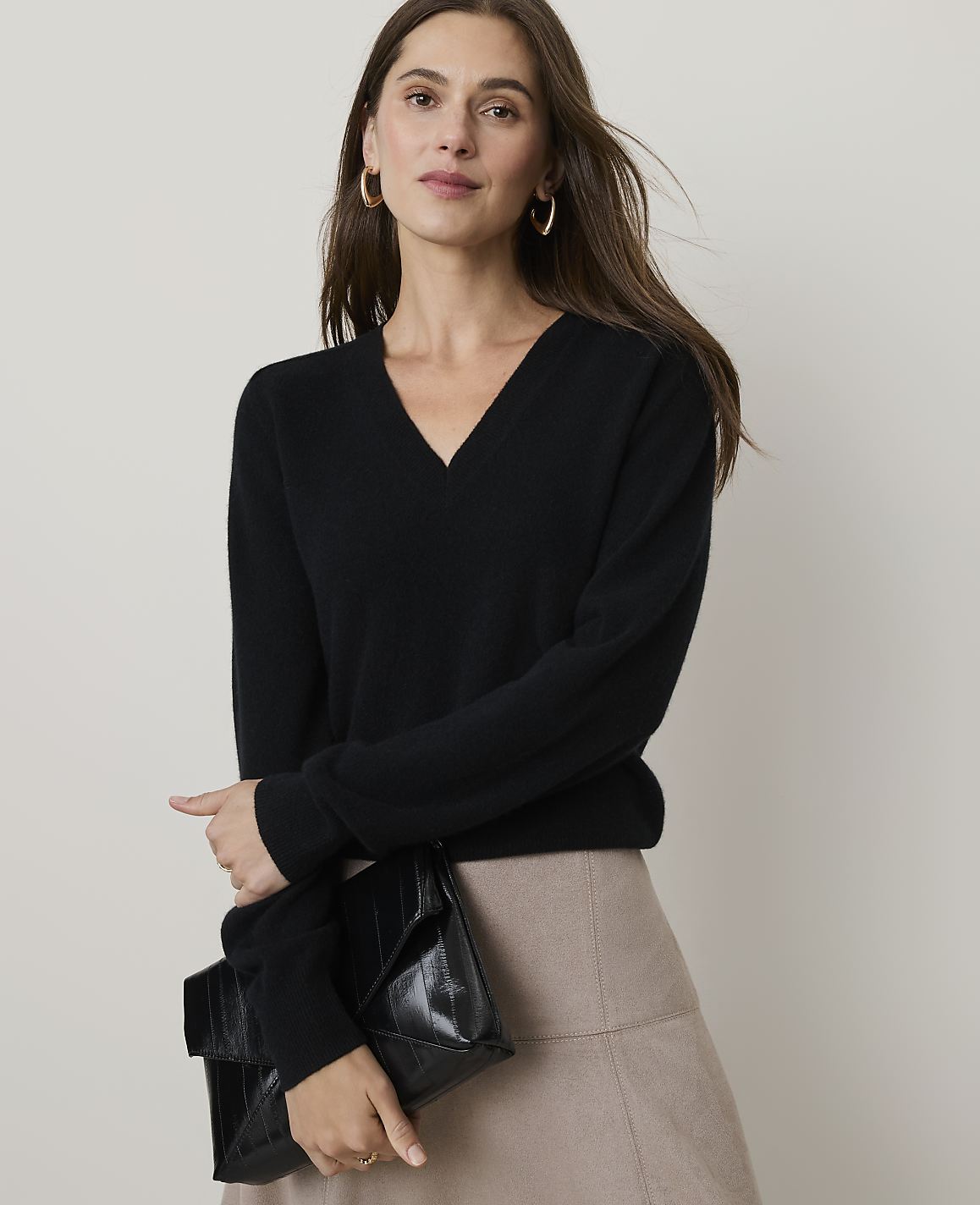 A woman in a black v-neck sweater and beige skirt holds a black clutch in front of a plain background.