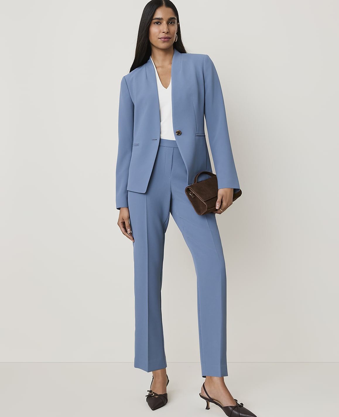 A woman in a blue suit sits on a stool, holding a woven bag, against a plain white background.