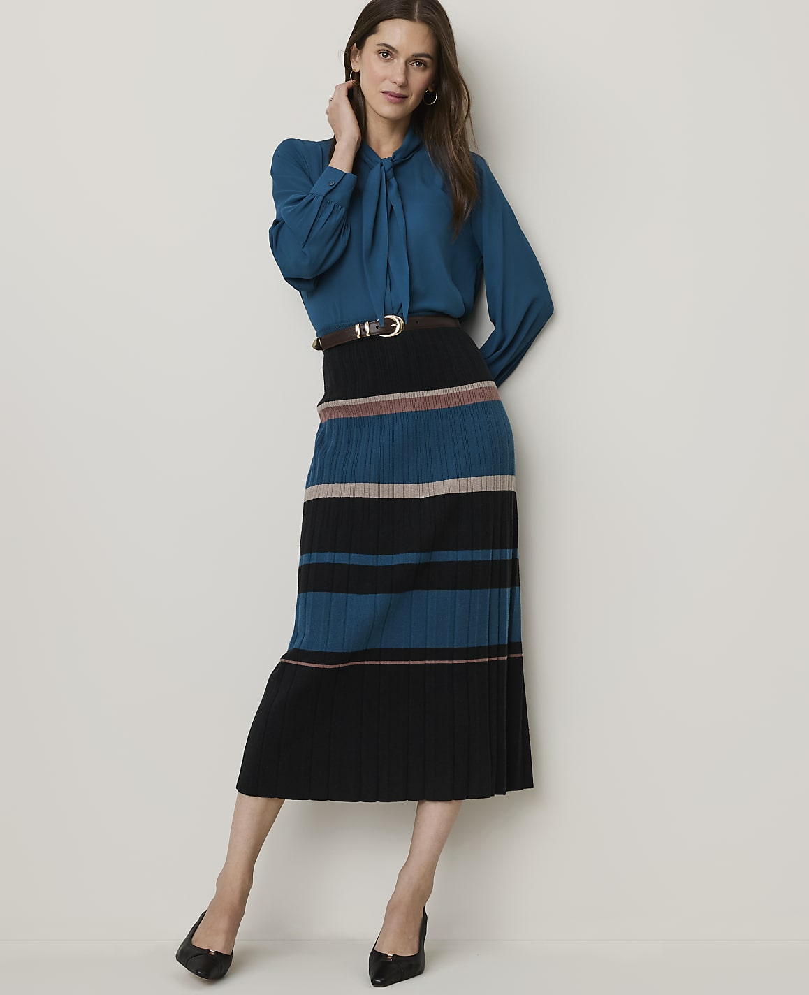 A woman in a teal blouse and a striped pleated skirt stands against a plain white background.