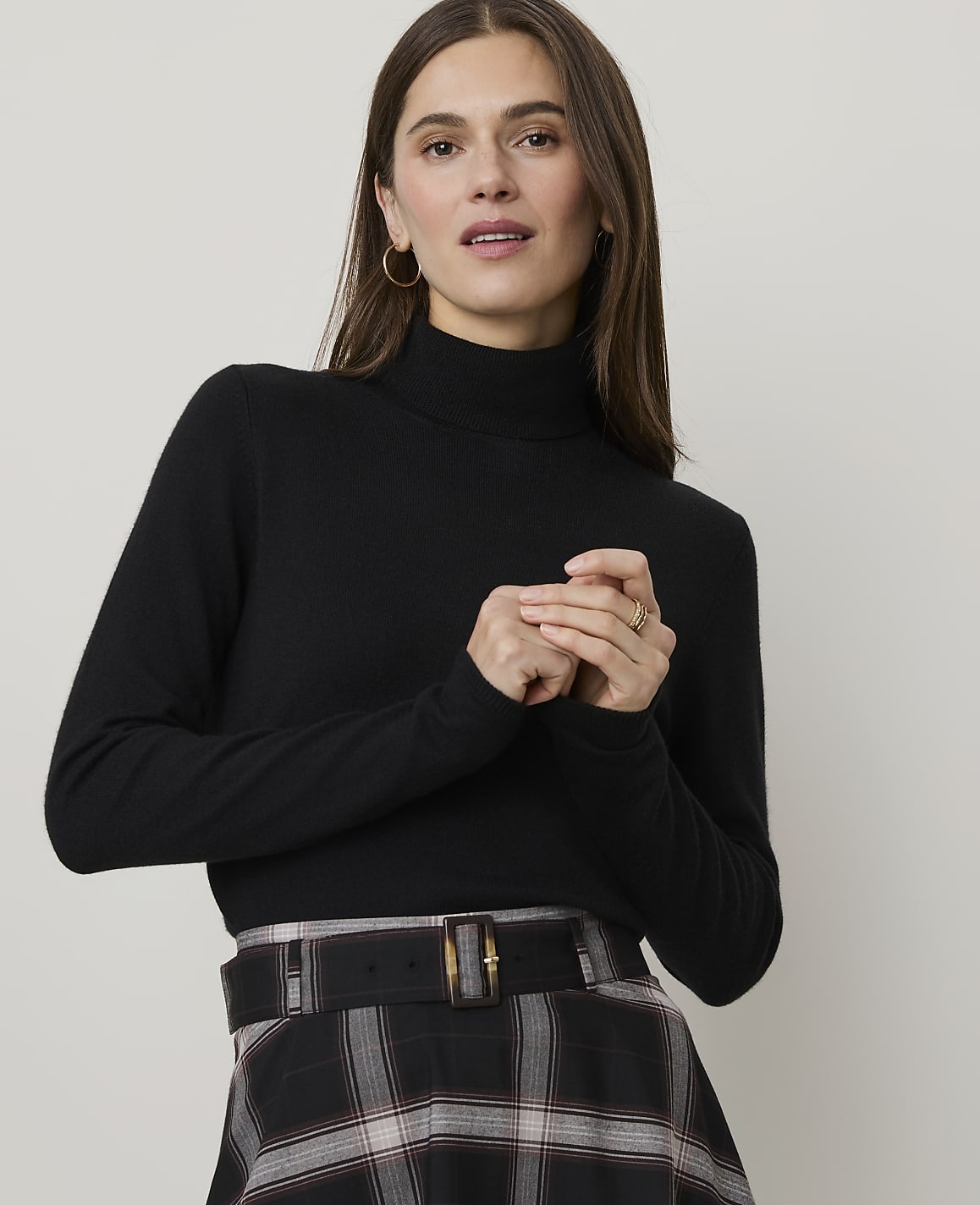 A woman in a black turtleneck and plaid skirt stands against a plain background.