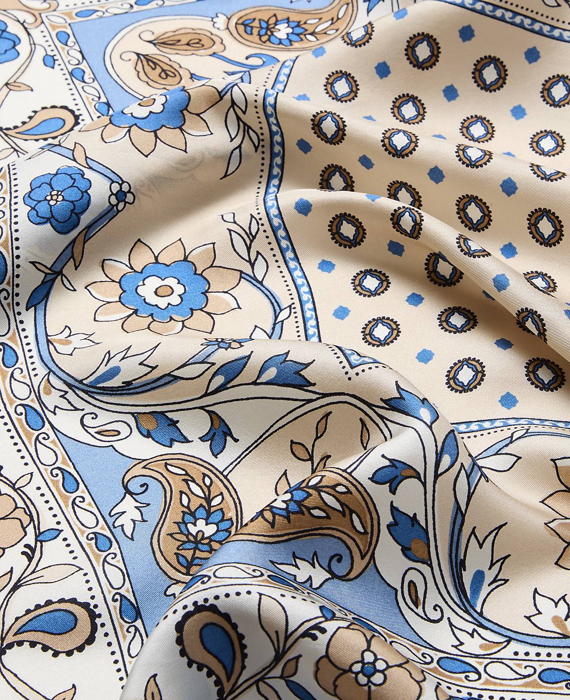 A close-up of a beige silk scarf with a blue and brown paisley and floral pattern in the foreground, set against a background of similar patterned fabric.