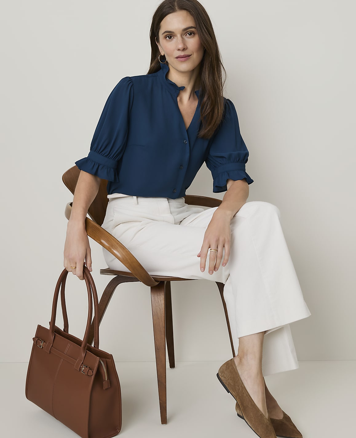 A woman in a teal blouse and white pants sits on a wooden chair holding a brown tote bag, with a plain, light-colored wall behind her.