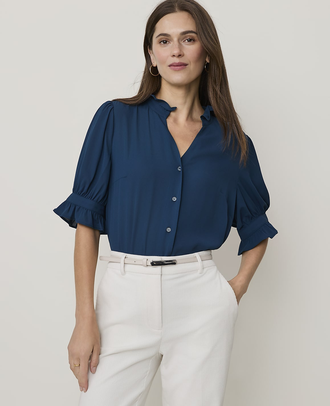 A woman in a teal blouse and cream pants stands against a plain white background.