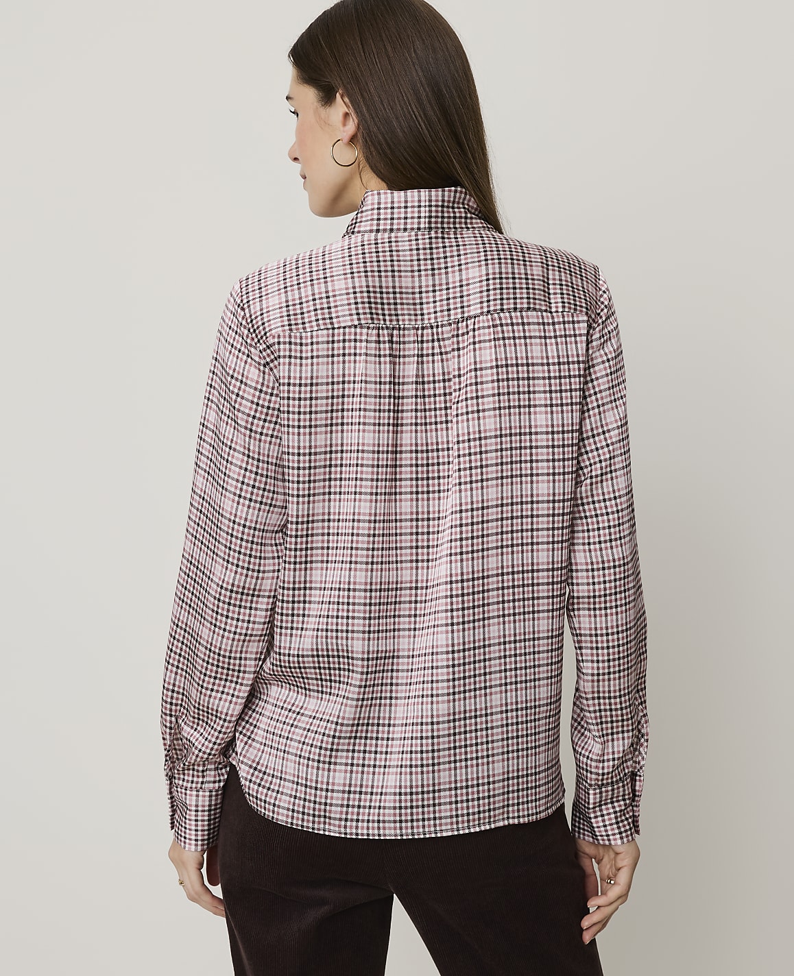 A woman wearing a pink plaid shirt and brown pants stands against a plain, light-colored background.