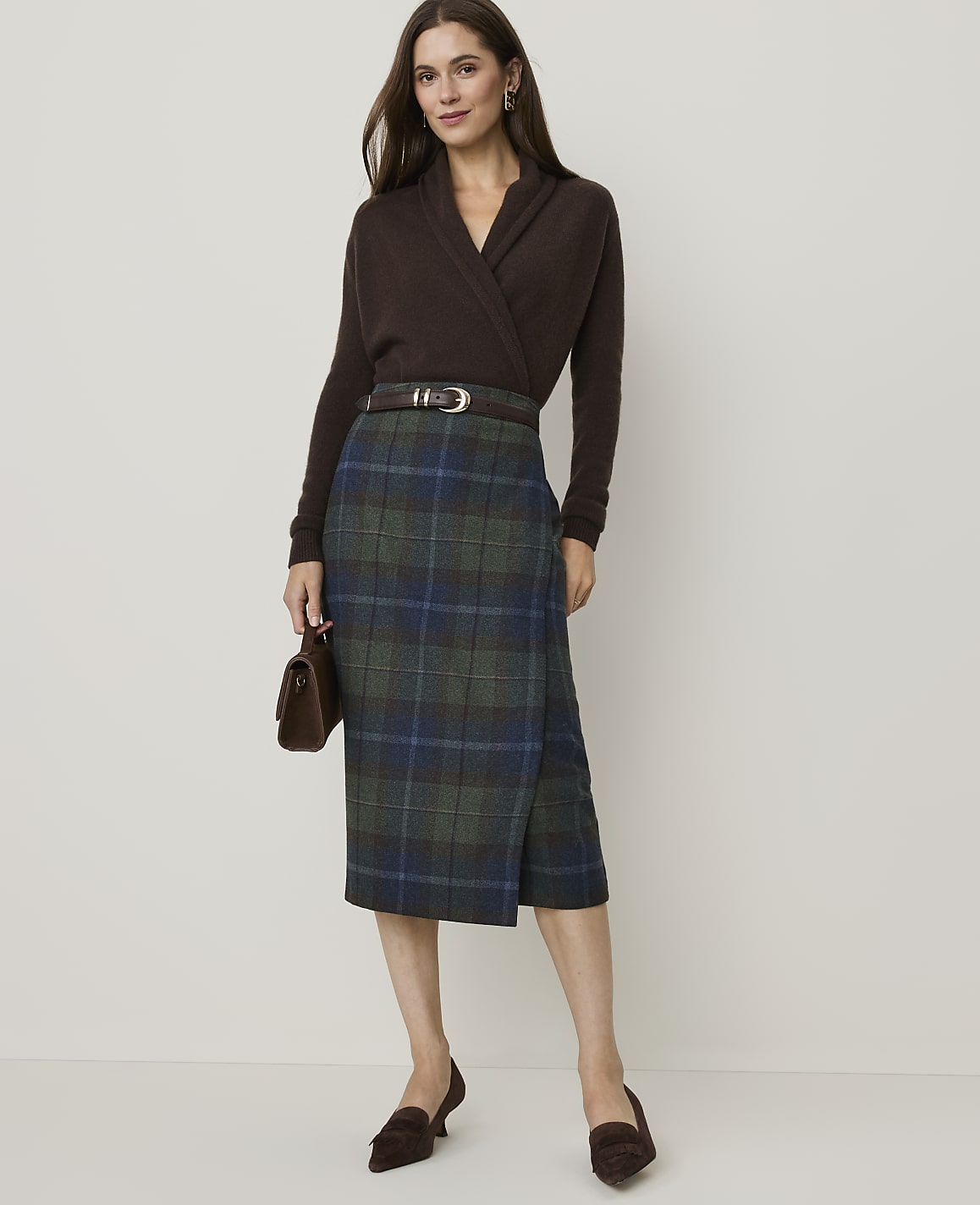 A woman in a brown sweater and plaid skirt stands against a plain background.