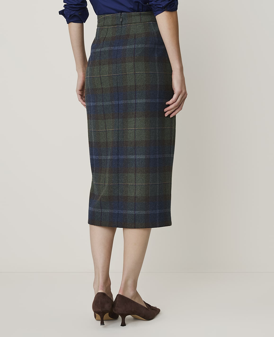 A woman in a plaid pencil skirt and brown heels stands against a plain, light-colored background.