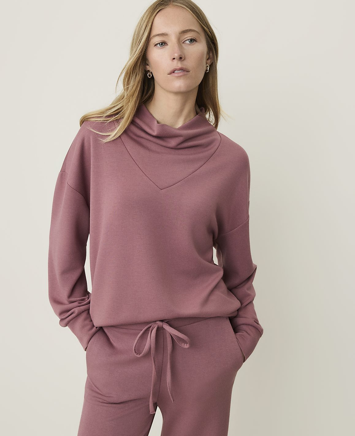 A woman in a rose-colored cowl neck sweatshirt and matching pants stands against a plain, light-colored background.