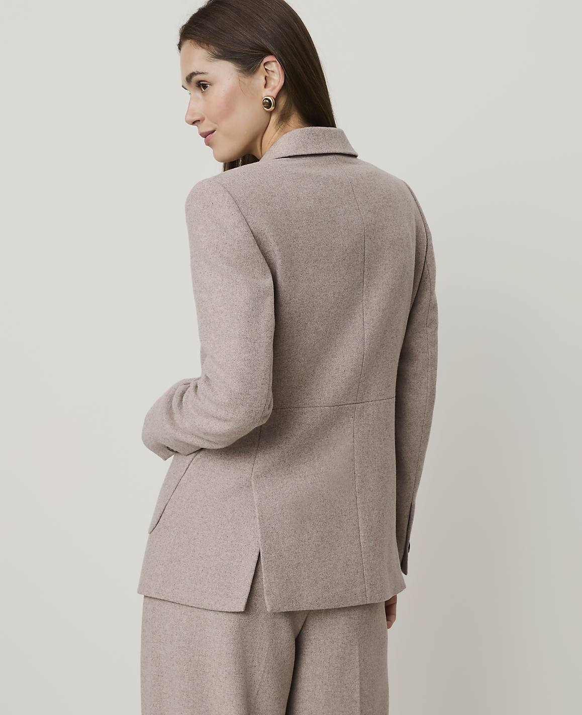 A woman in a beige blazer and pants stands against a plain, light-colored background.