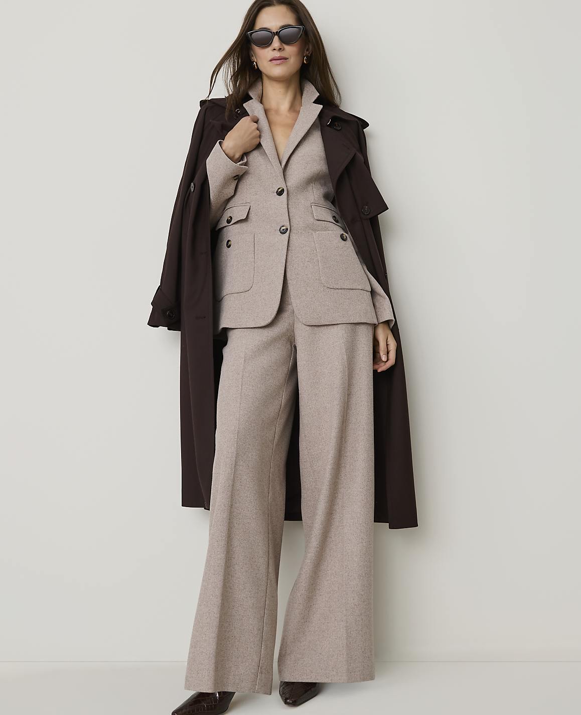 A woman in a beige suit and brown trench coat stands against a plain white background.