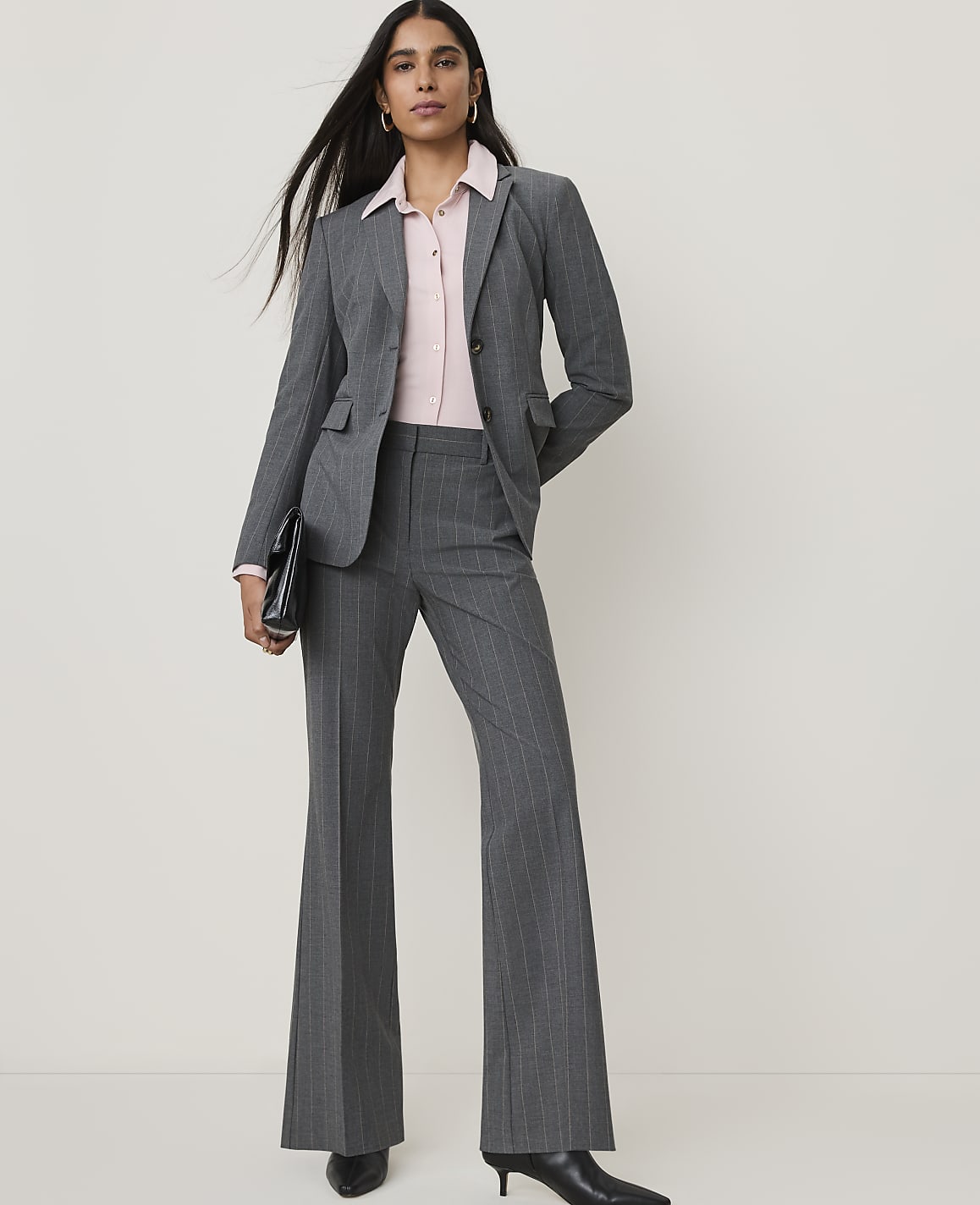 A woman in a gray pinstripe suit and pink blouse stands against a plain white background, holding a black clutch.
