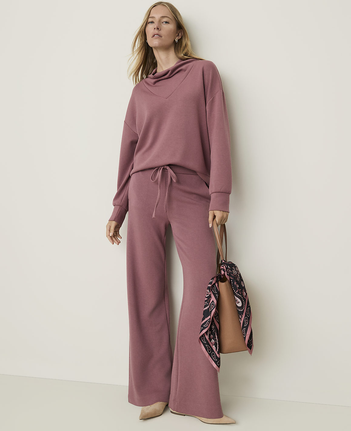 A woman in a mauve loungewear set stands against a plain, light-colored wall, holding a brown tote bag with a patterned scarf.