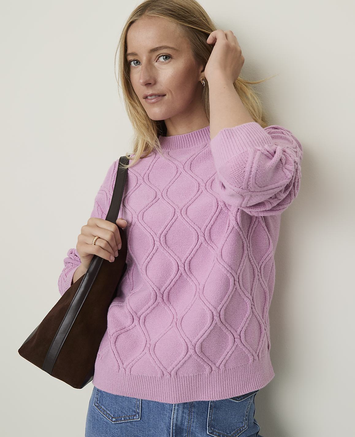 A woman in a pink cable-knit sweater and jeans stands against a plain, light-colored wall, with a brown handbag slung over her shoulder.