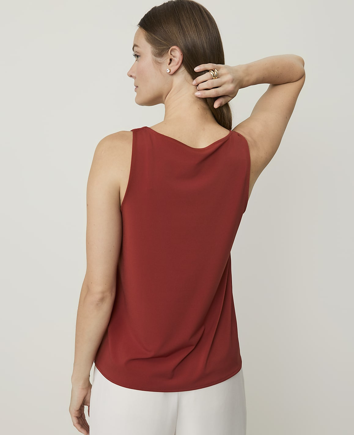 A woman in a rust-colored tank top and white pants stands against a plain, light background, with her hand behind her head.