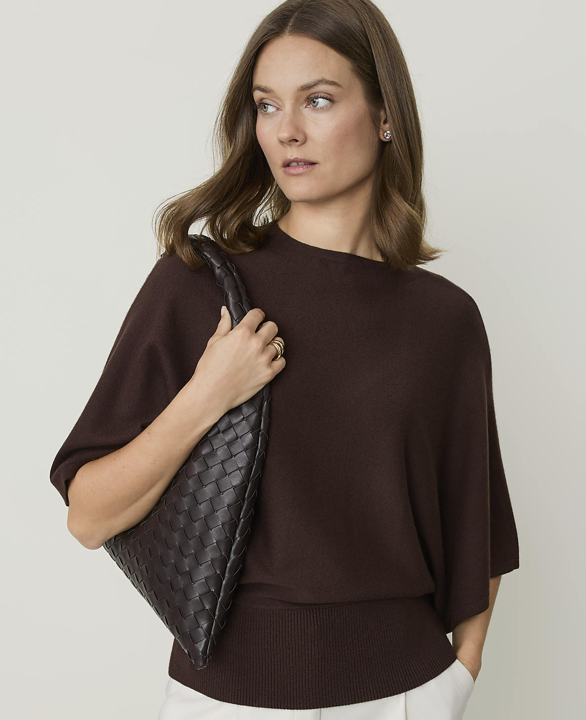 A woman in a brown sweater and white pants holds a woven brown purse against a plain background.