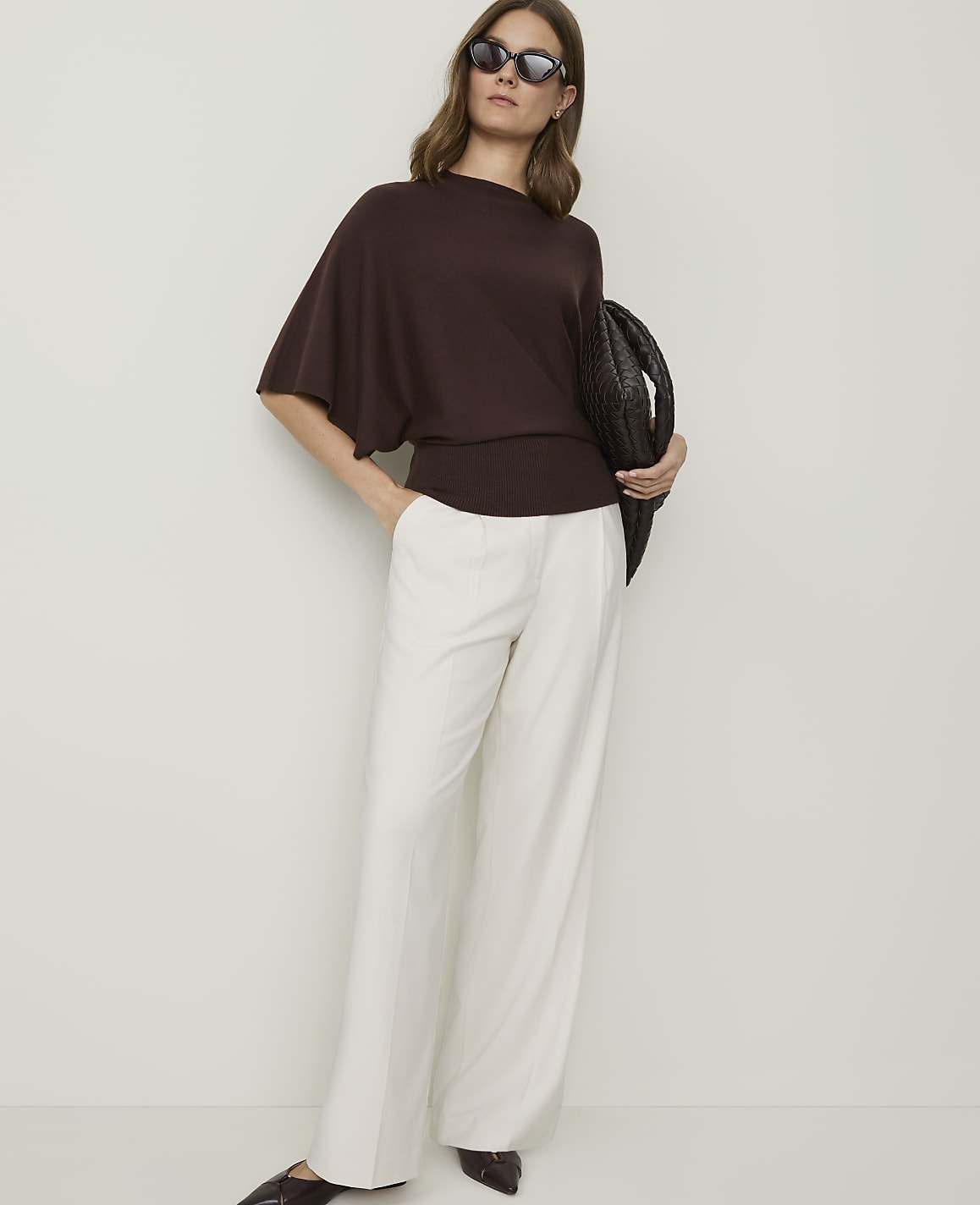 A woman in a brown sweater and white pants stands against a plain background, holding a woven bag.