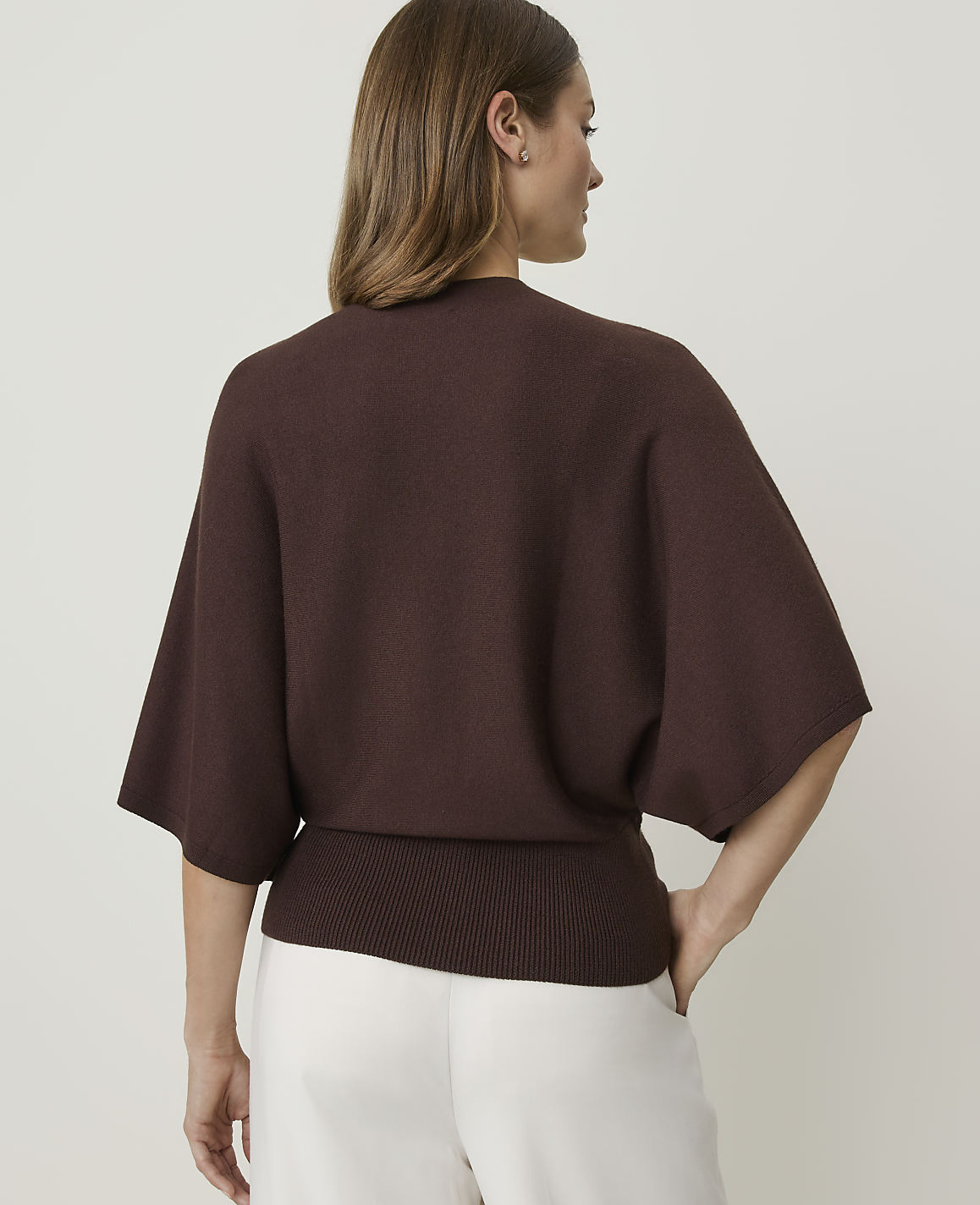 A woman in a brown sweater and white pants stands against a plain white background.