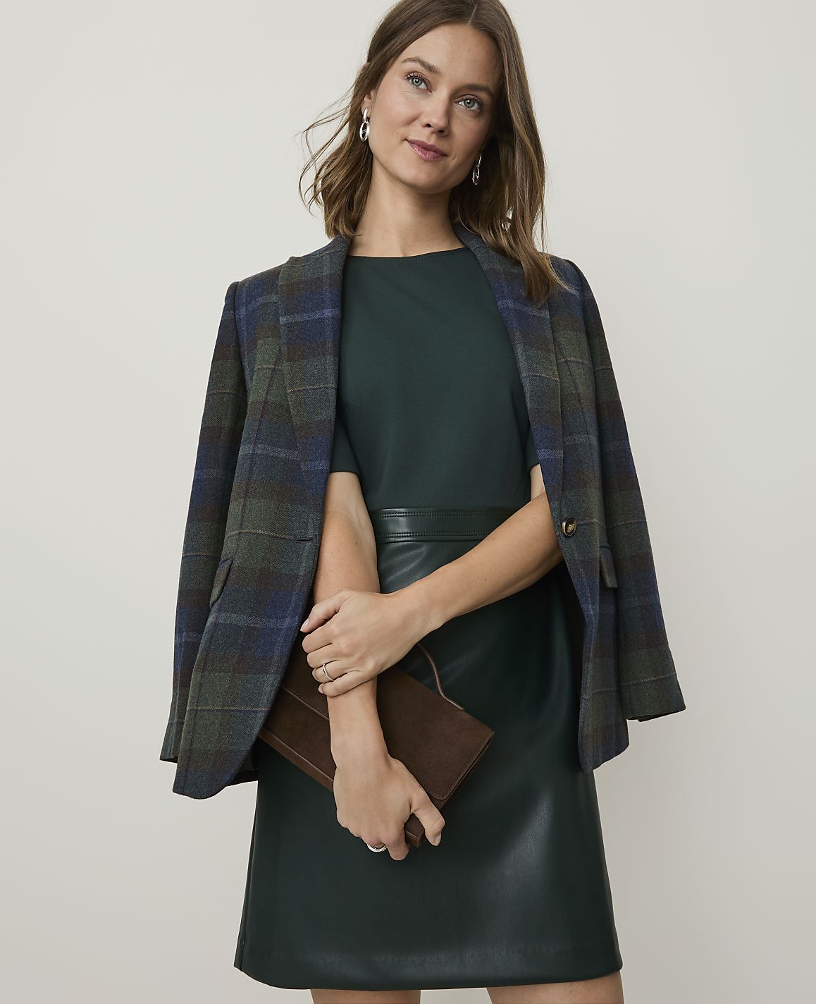 A woman in a green dress and plaid blazer holds a brown clutch in front of a plain background.
