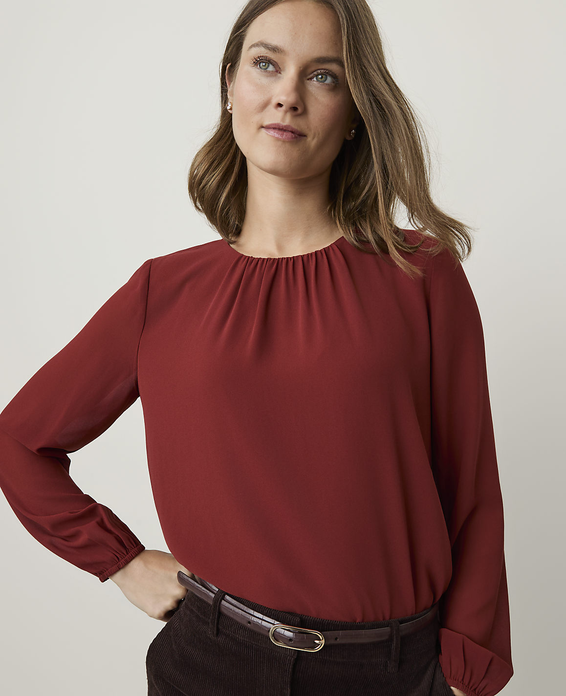 A woman in a rust-colored blouse and dark pants stands against a plain, light background.