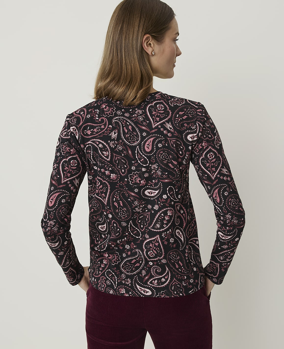 A woman in a black paisley-patterned shirt stands with her hands in her pockets against a plain, light-colored background.