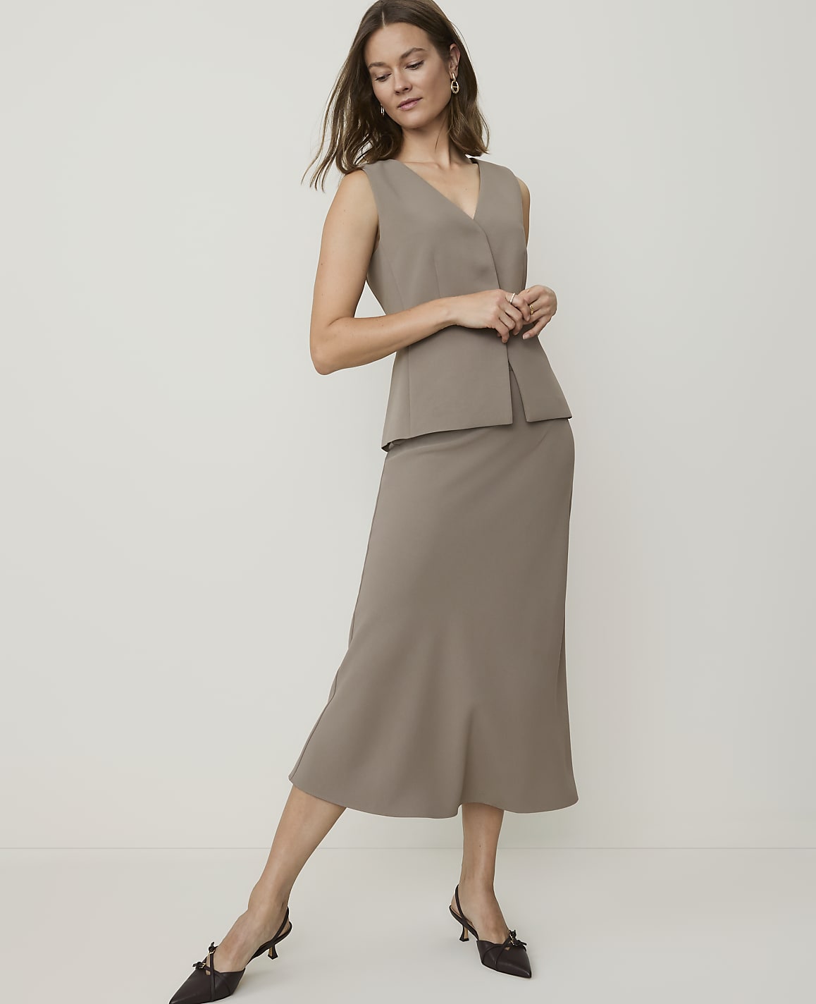 A woman in a taupe sleeveless top and matching midi skirt poses against a plain, light-colored background.