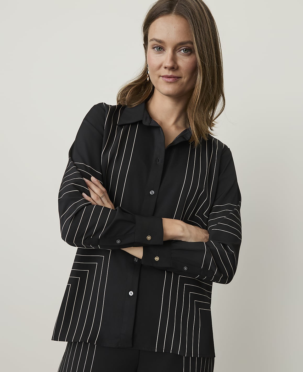 A woman in a black shirt with white geometric patterns stands with her arms crossed against a plain, light gray background.