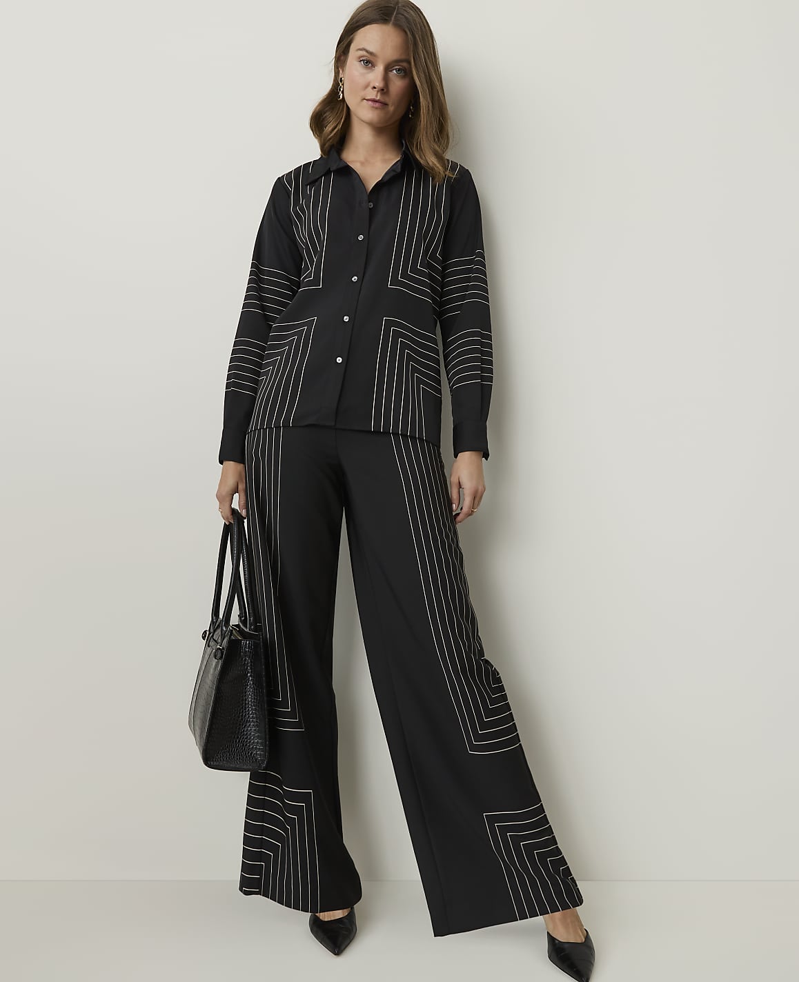 A woman in a black patterned shirt and wide-leg pants stands against a plain white background, holding a black handbag.