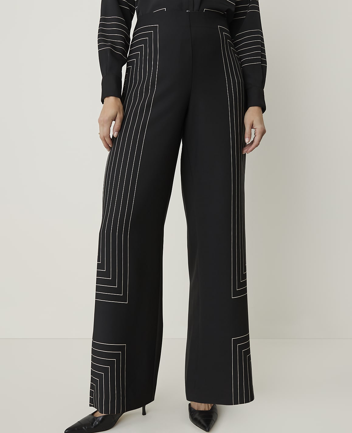A person stands in wide-leg black pants with geometric white stitching, against a plain white background.
