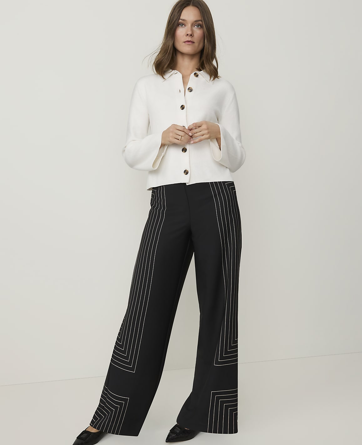 A woman in a white jacket and black patterned pants stands against a plain white background.