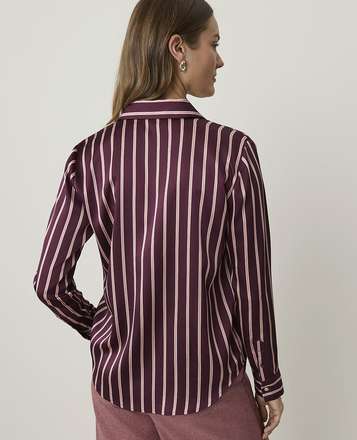 A woman in a burgundy and pink striped satin shirt stands against a plain, light-colored background.