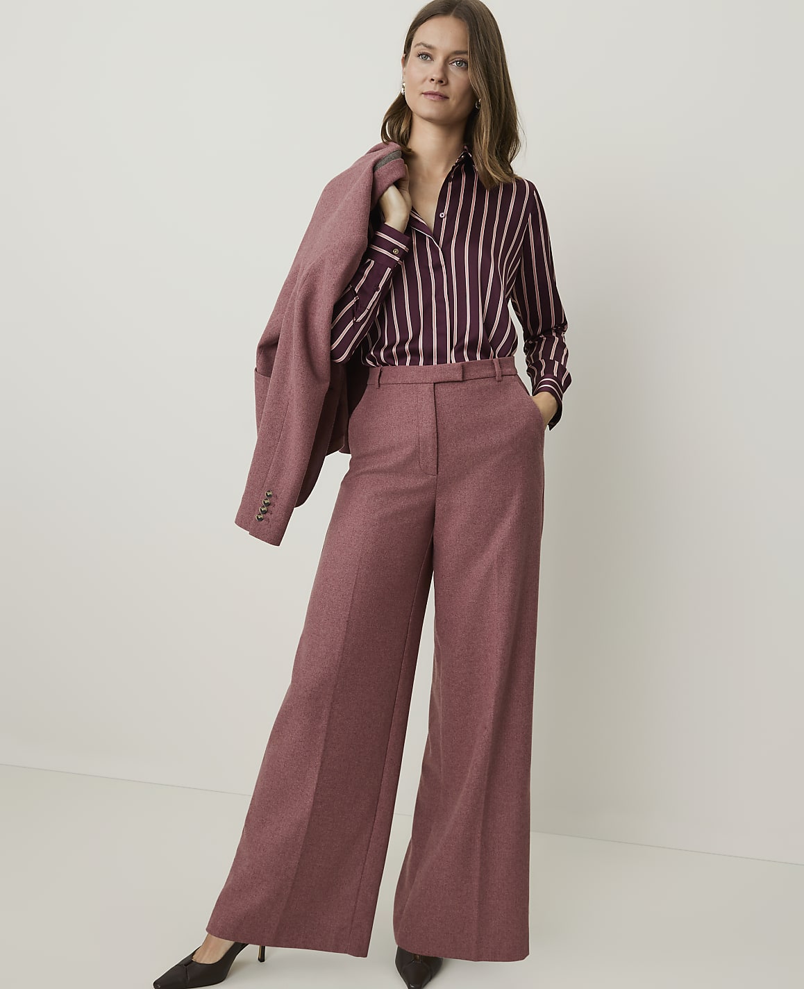 A woman in a striped blouse and wide-leg pants stands against a plain, light-colored background, with a blazer draped over her shoulder.
