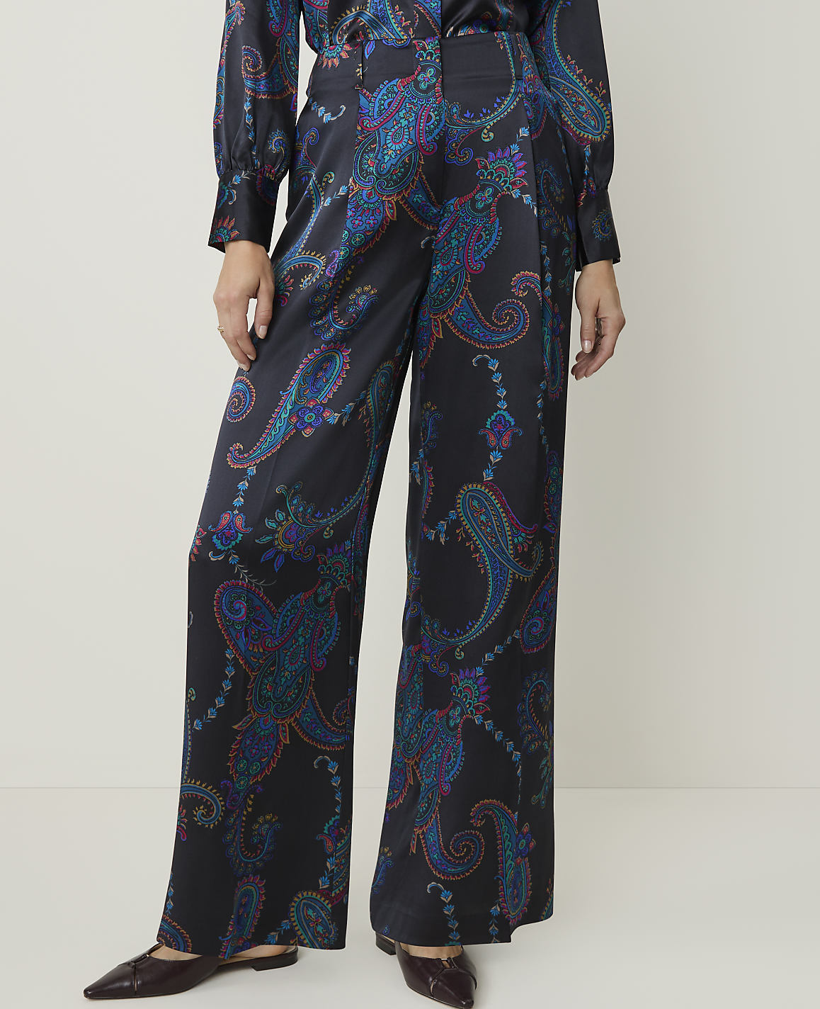 A person stands in wide-leg, dark satin pants with a colorful paisley pattern, against a plain, light-colored background.