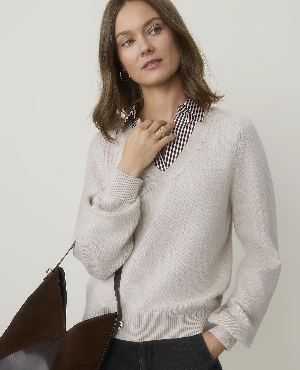 A woman in a cream v-neck sweater over a striped shirt stands against a plain, light background, with a brown bag in the foreground.