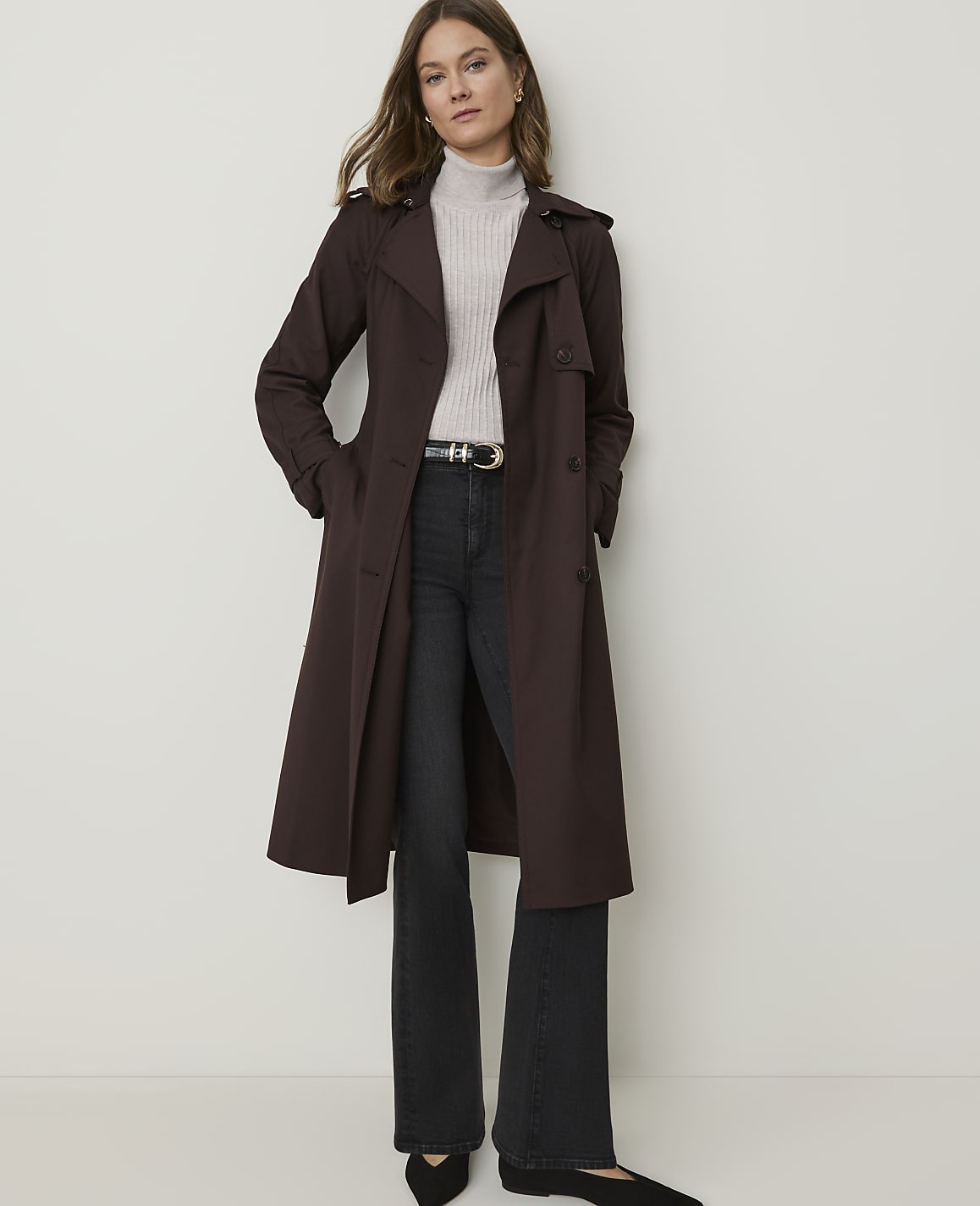 A woman in a brown trench coat and black flared jeans stands against a plain white background.