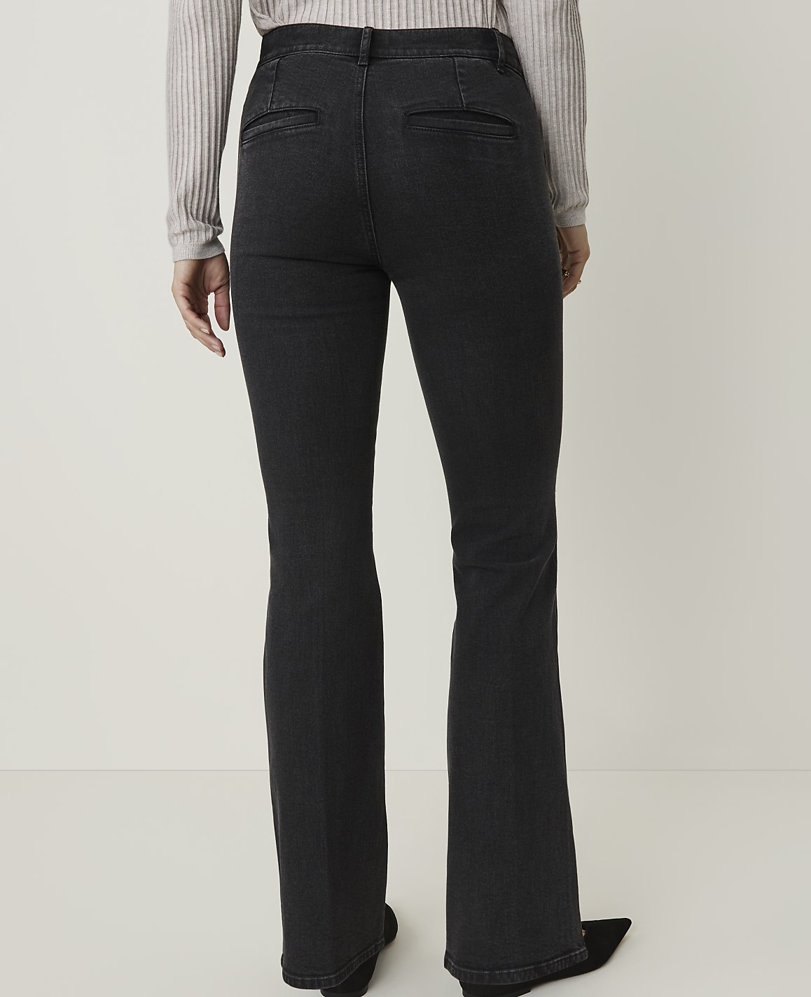 A person wearing dark wash flared jeans and a light grey sweater stands against a plain white background.