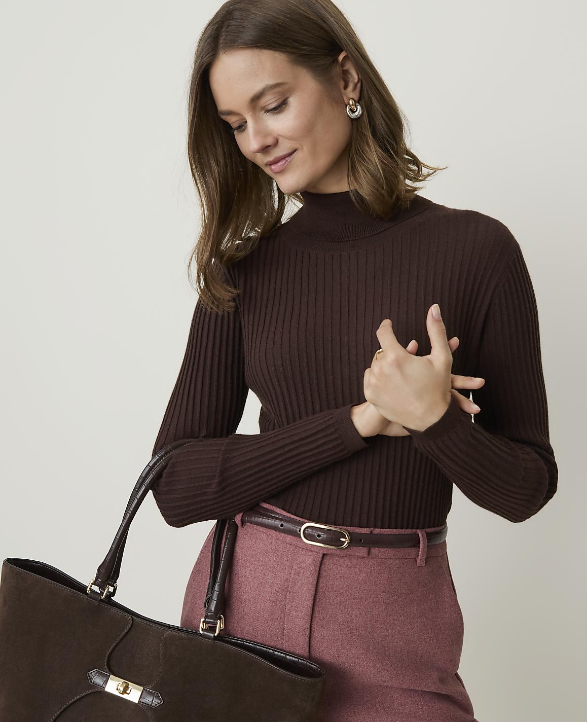 A woman in a brown turtleneck and pink pants holds a brown suede tote bag against a plain background.
