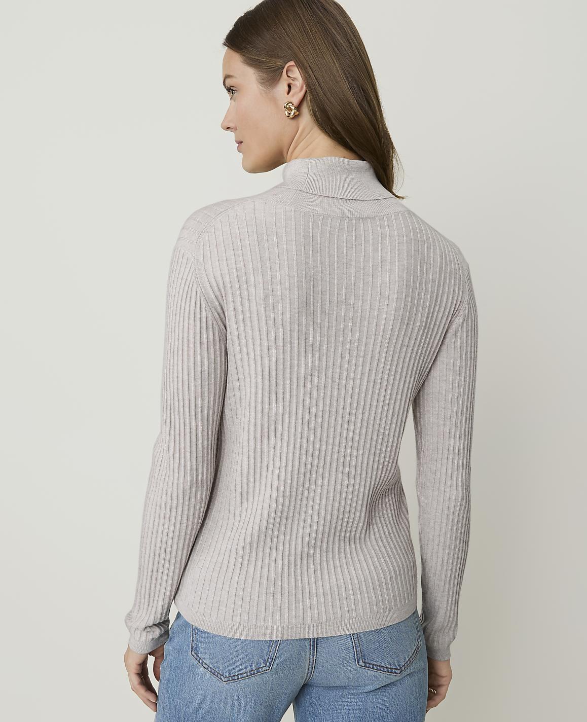 A woman in a light gray ribbed turtleneck sweater and blue jeans stands against a plain, light gray background.