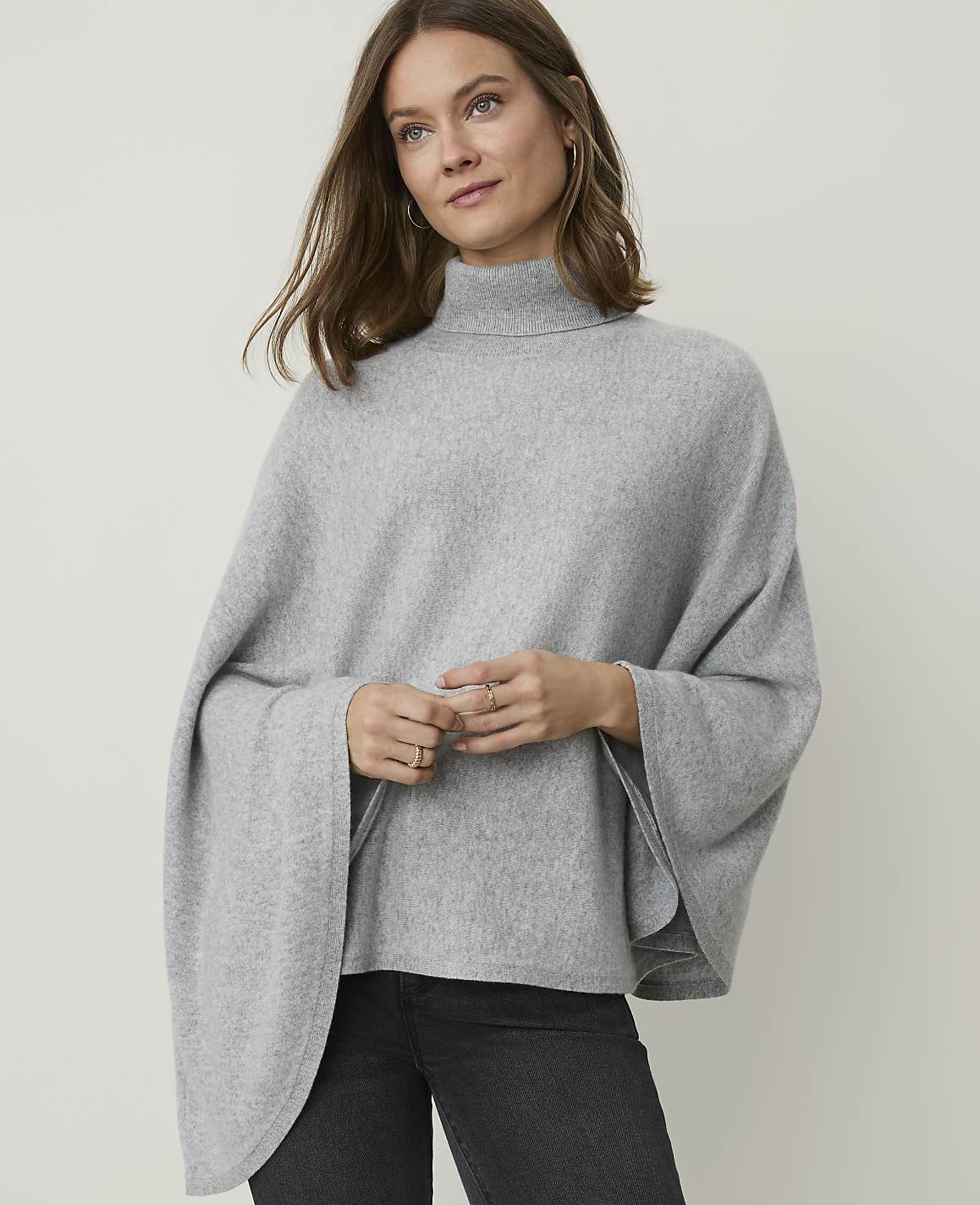 A woman in a grey turtleneck poncho stands against a plain grey background.
