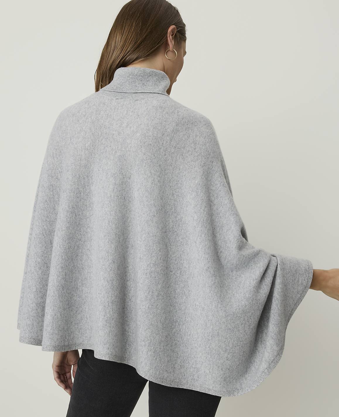 A woman wearing a light gray turtleneck poncho stands against a plain, light gray background.