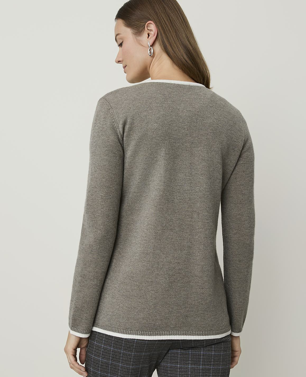 A woman in a taupe sweater with white trim stands against a plain, light gray background.