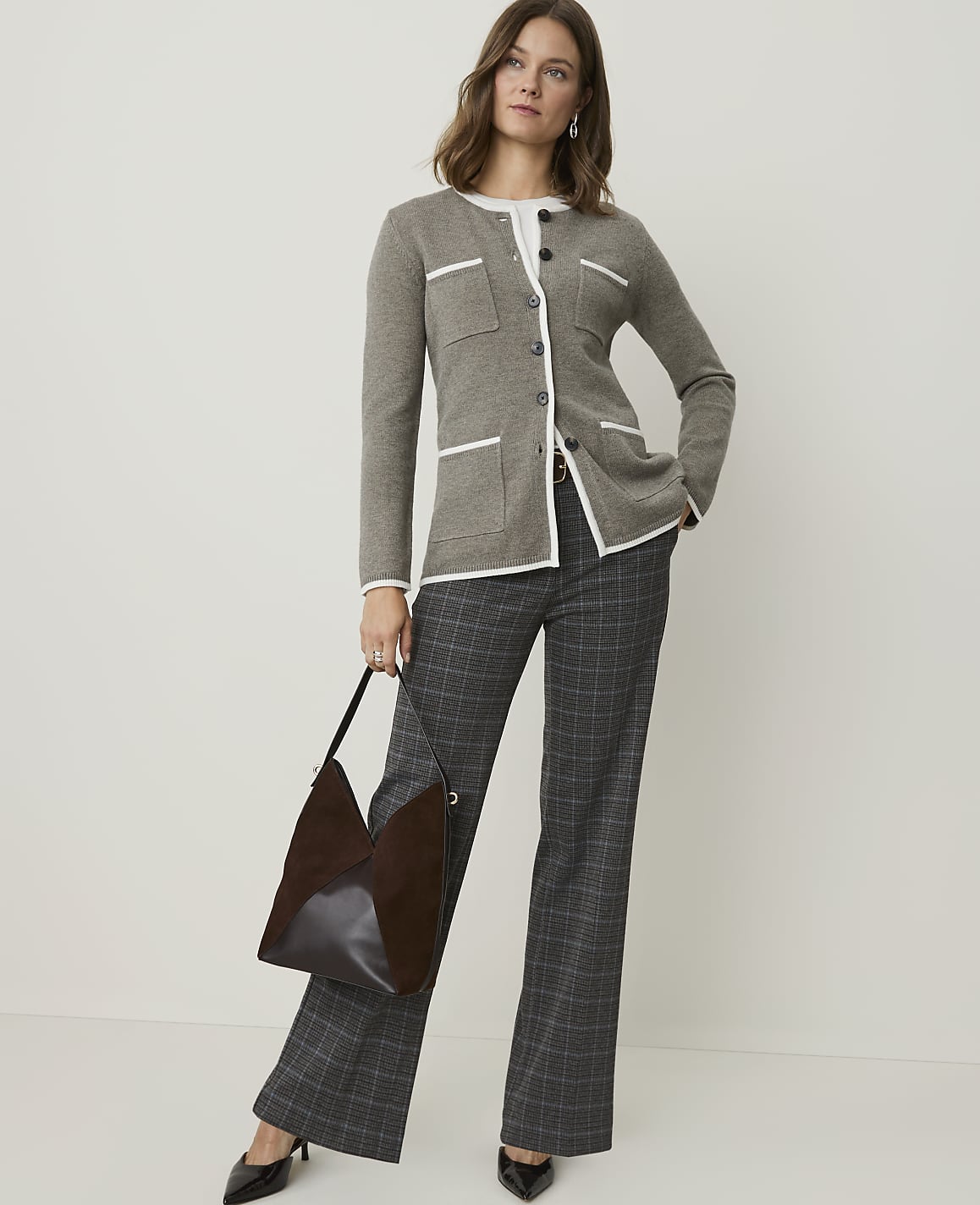 A woman in a grey cardigan and plaid pants stands against a plain background, holding a brown handbag.