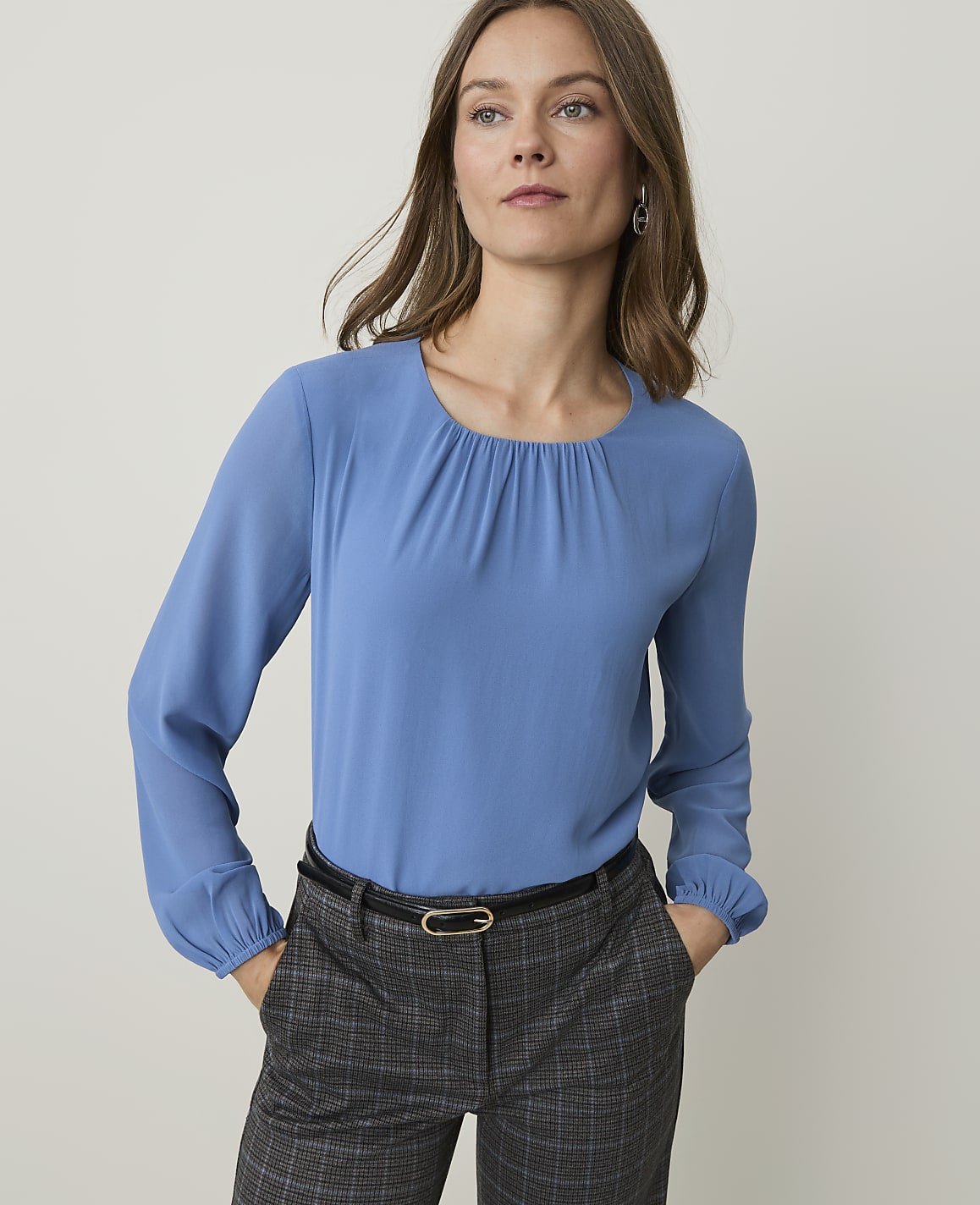 A woman in a blue blouse and plaid pants stands against a plain background.