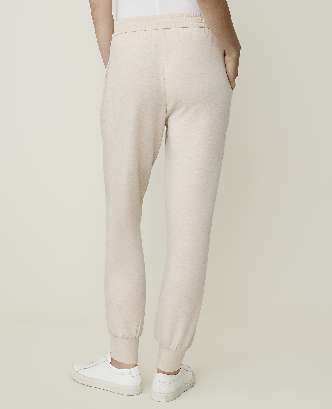 A person wearing light-colored sweatpants and white sneakers stands against a plain, light-colored background.