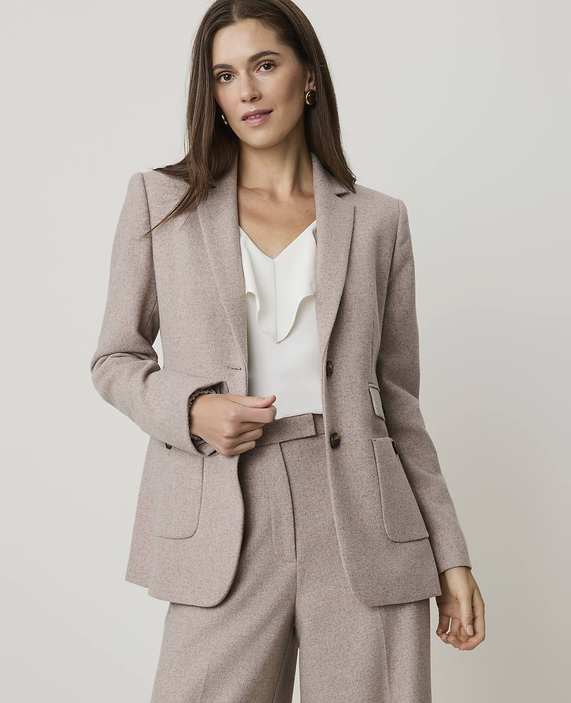 A woman in a beige blazer and pants stands against a plain gray background.