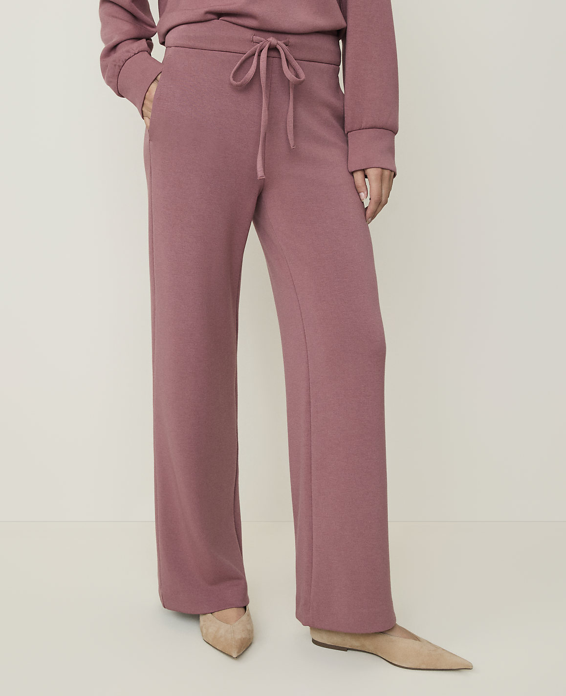 A person wearing rose-colored lounge pants and beige flats stands against a plain, light-colored background.