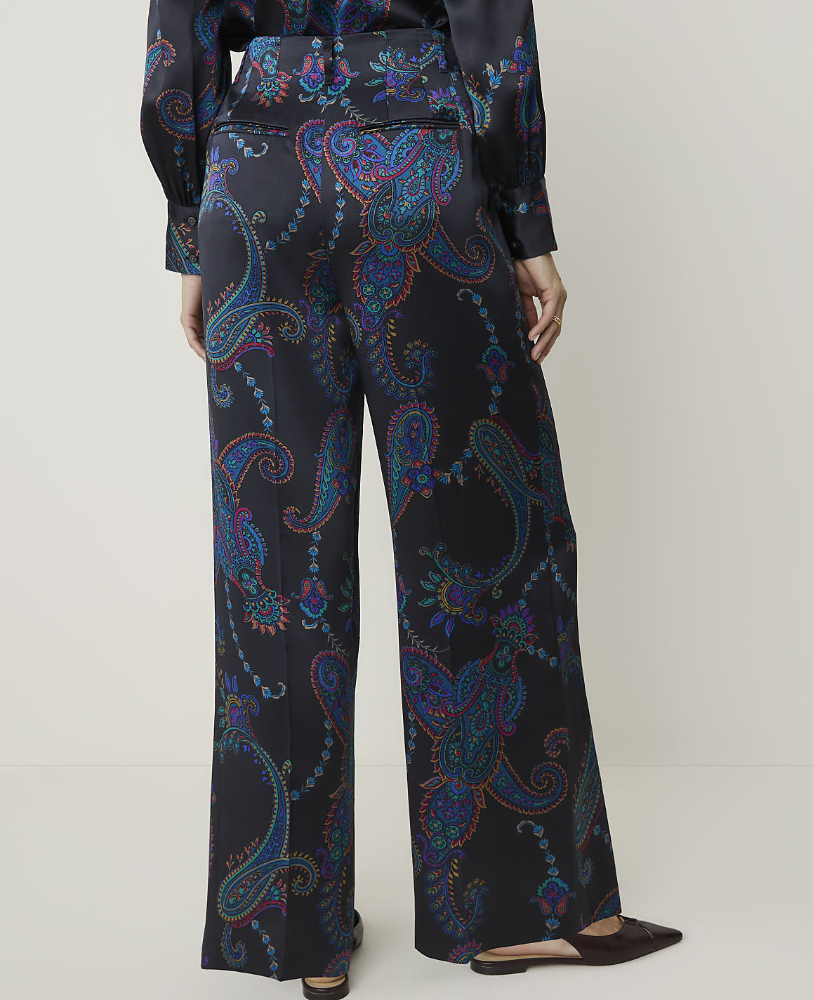A person stands in wide-leg paisley-patterned pants and dark flats in the foreground, against a plain, light-colored background.