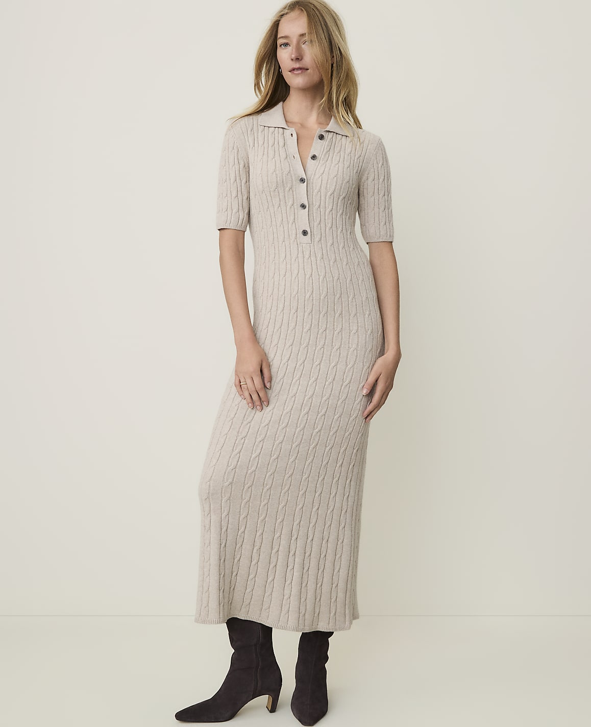 A woman in a beige cable-knit midi dress and dark boots stands against a plain, light-colored background.