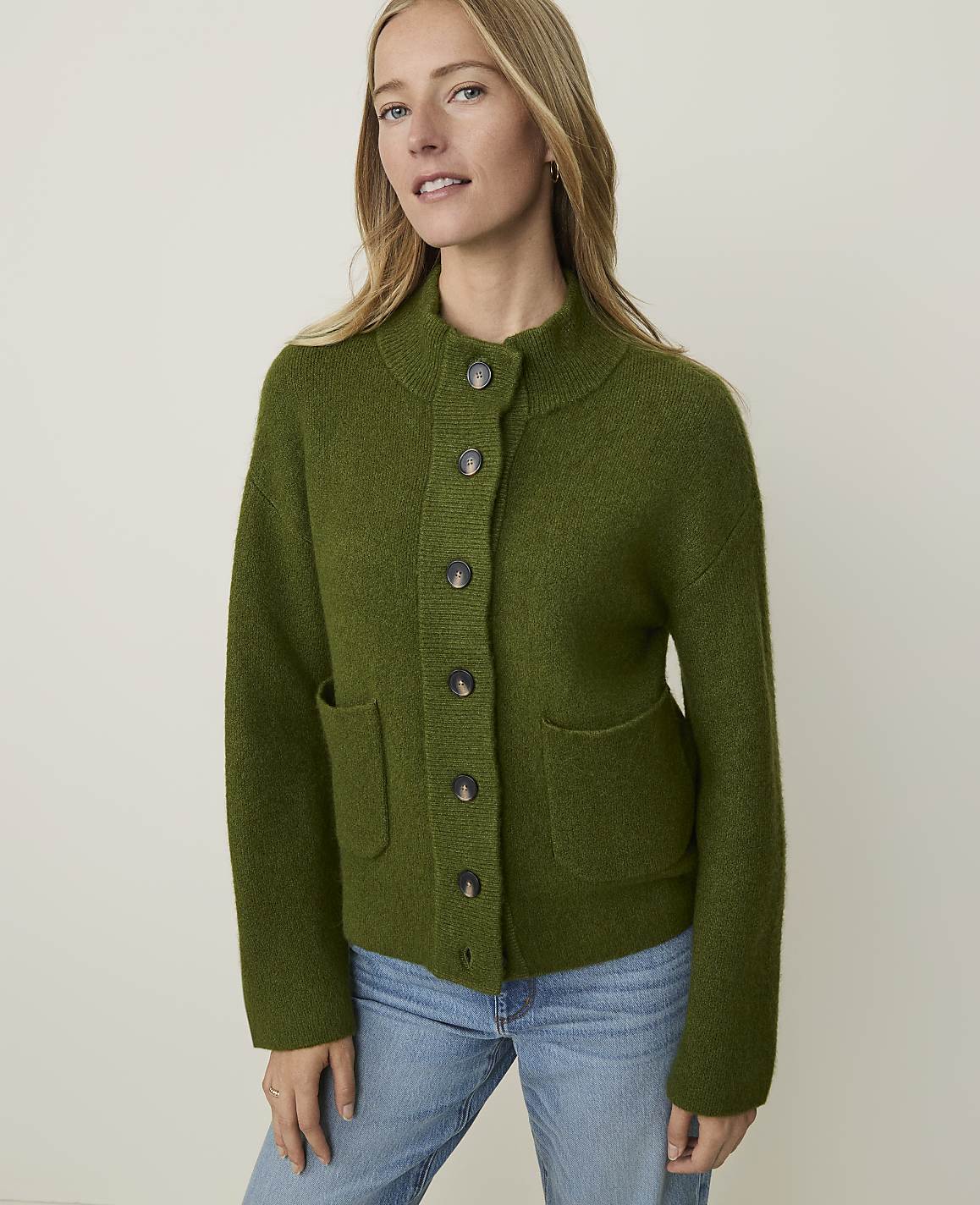 A woman in a green cardigan and jeans stands against a plain, light background.